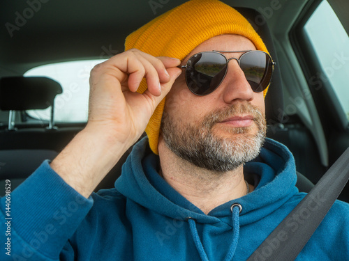 Portrait of a man driver in blue jacket and yellow hat wearing dark aviator glasses. Travel and transport. Male model in his 50s with grey beard dressed for outdoor adventure.