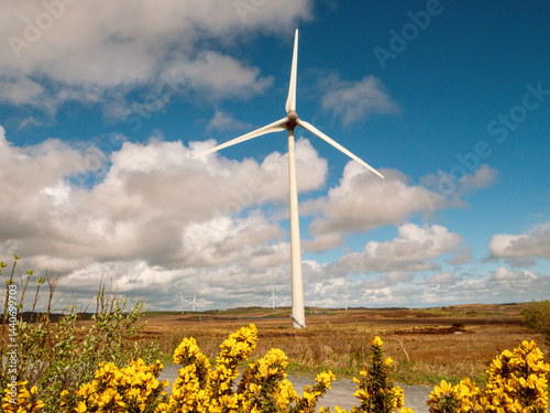 A field of wind turbines with a clear blue cloudy sky in the background. The windmills are tall and spread out across the field. Production of clean green energy. Warm sunny day. Nobody.