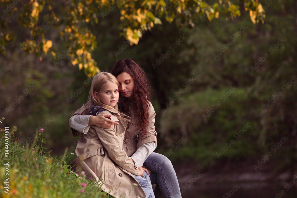 Lifestyle photo of mother with daughter spending time together sitting on grass in park. Curly woman hugs blonde girl outdoors. Family value and support.