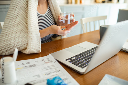Sick woman taking medication while working from home with laptop and bills on kitchen table