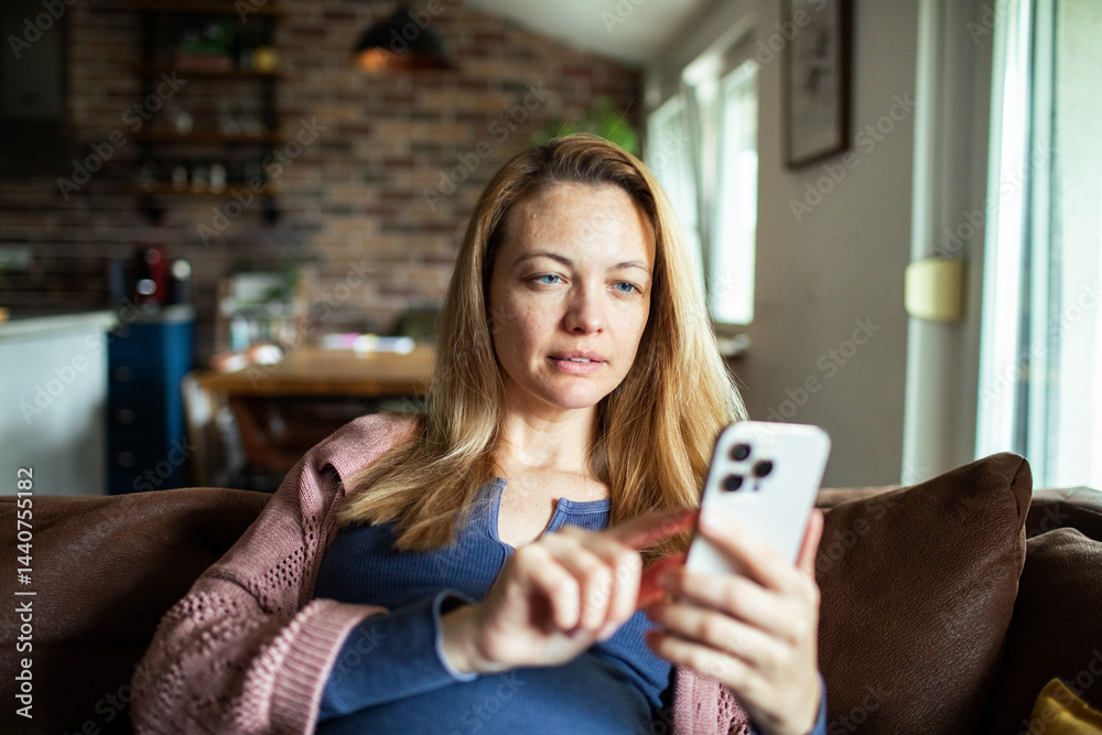 Pregnant woman using mobile banking app on smartphone while relaxing on couch