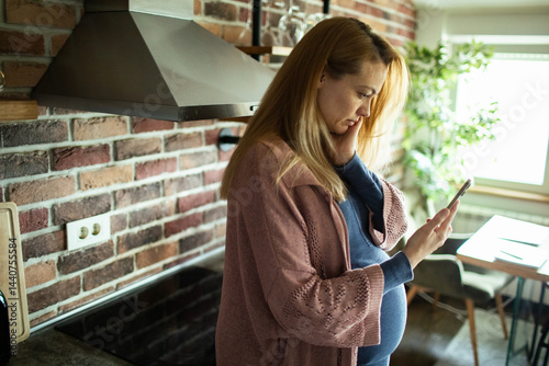 Pregnant woman reading phone message in modern kitchen