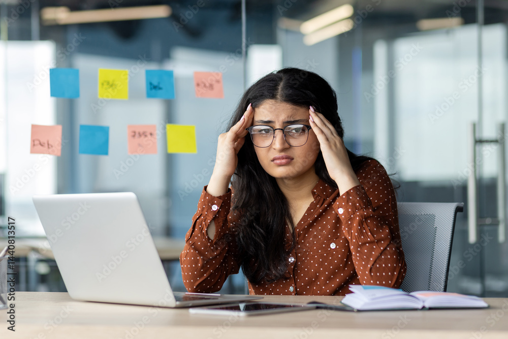 A young woman looks stressed while working on her laptop in an office environment. She appears overwhelmed by the task at hand.