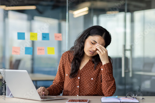 A businesswoman experiencing stress while working on her laptop at her desk in an office setting. She is rubbing her eyes.
