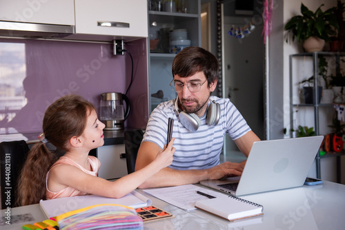 Father is working from home, wearing headphones around his neck, while his daughter is trying to get his attention by showing him some colored pencils