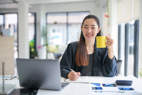Asian businesswoman drinking coffee and working with laptop and taking notes on notebook in modern office