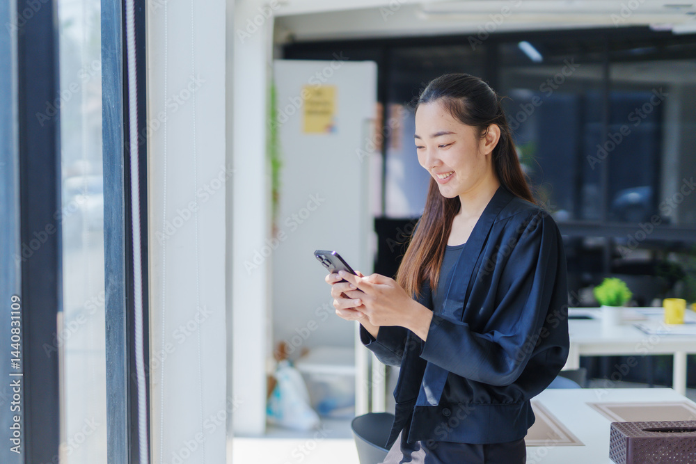 Smiling businesswoman using a smartphone while standing near a bright window in a modern office, enjoying a productive workday Smiling businesswoman using a smartphone while standing near a bright window in a modern office, enjoying a productive workday