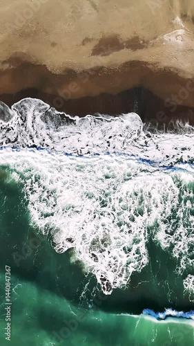 Aerial view capturing emerald green waves breaking over golden sand, white foam cascading along pristine coastal landscape with serene marine scenery