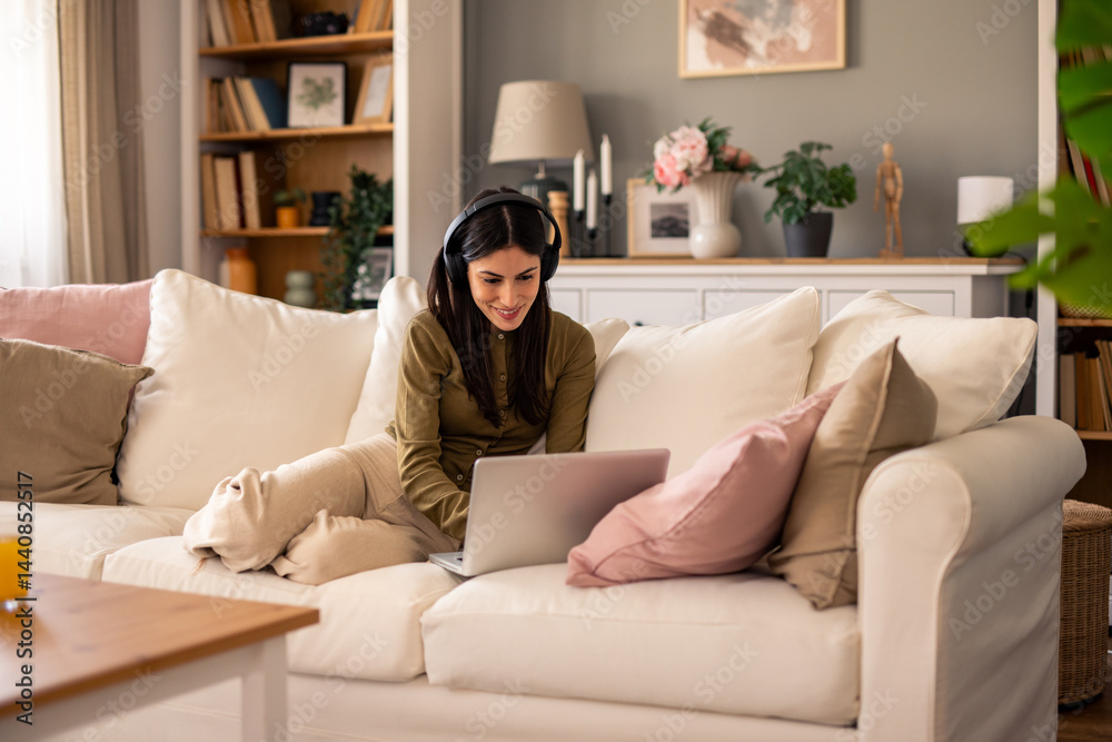 Young Woman Wearing Headphones Using Laptop in a Cozy Living Room with Cushions and Decor Young Woman Wearing Headphones Using Laptop in a Cozy Living Room with Cushions and Decor