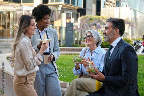 Multi ethnic colleagues eating healthy food and talking during lunch break in a green area of the business district