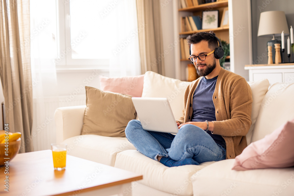 Man Wearing Headphones on Couch Working on Laptop in a Bright Comfortable Living Room