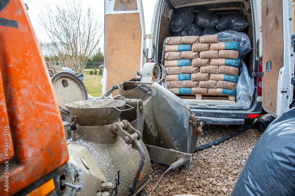 Outdoor construction site featuring a van with open rear doors, cement bags on a pallet, a concrete mixer, and tools on a gravel surface near leafless trees.