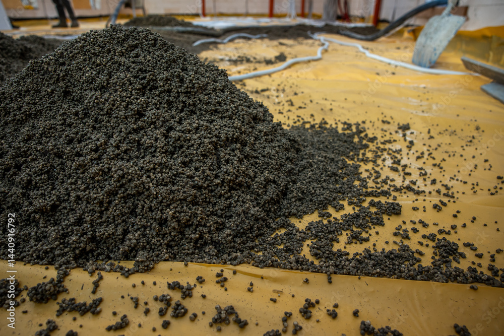 Dark granular material spread on a yellow surface in an indoor industrial facility, with visible pipes, hoses, and equipment in the background.