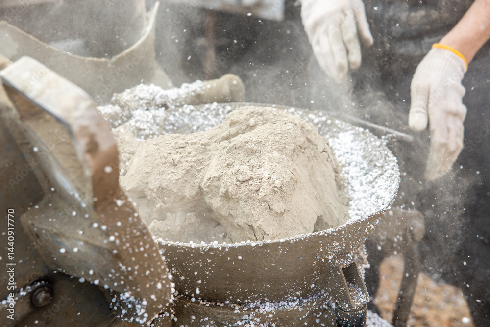 Powdered material and white granules mix in a metal container, with gloved hands nearby. Dust particles are visible, indicating an industrial process.