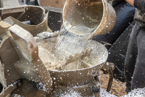 Water pours from a metal bucket into a concrete mixer, with splashes and droplets mid motion. Gravel ground and industrial equipment suggest construction work.