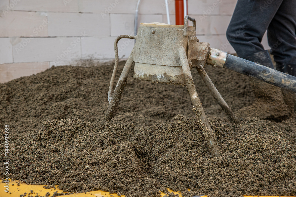 A concrete screed tool with a tripod structure and hose rests on lightweight aggregate concrete. A worker stands nearby near a white brick wall and red pipe.