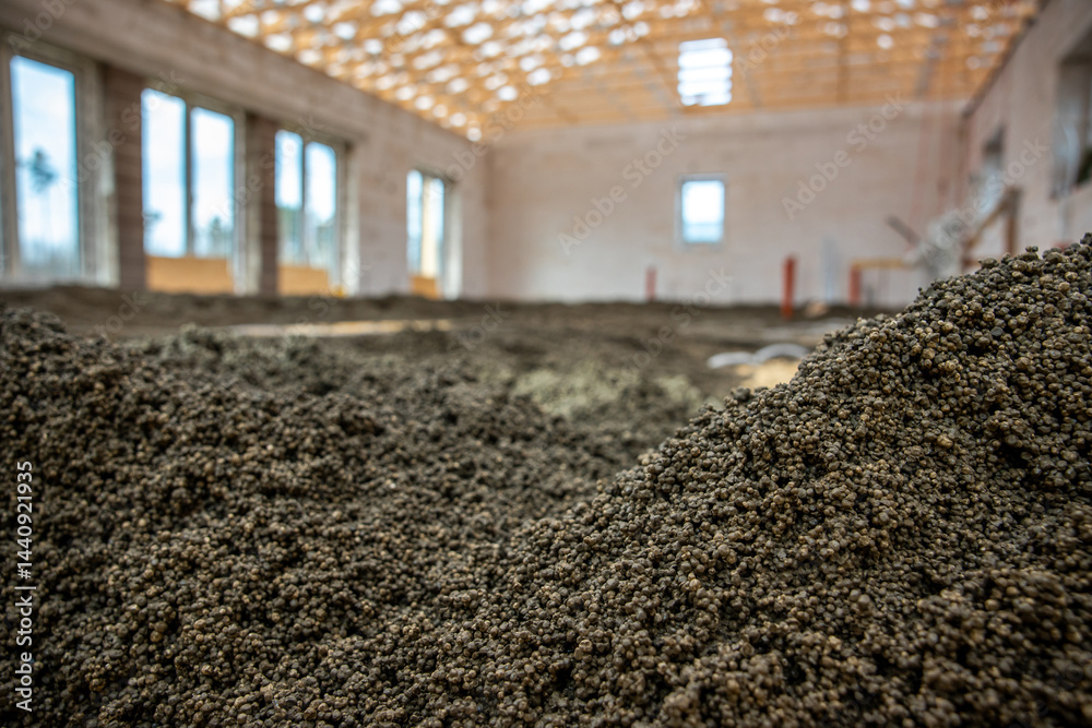 Construction site showing an exposed wooden roof framework, unfinished walls with large window openings, and granular material spread on the floor.