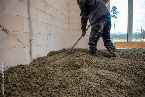Obraz na płótnie A worker uses a long handled tool to level wet concrete inside a building with white block walls. A large window reveals sparse trees and an open field. w salonie