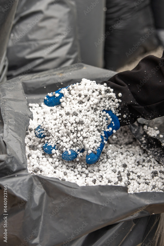 A blue and black gloved hand holds white and gray granules from an open plastic bag, with additional bags visible in an industrial setting.