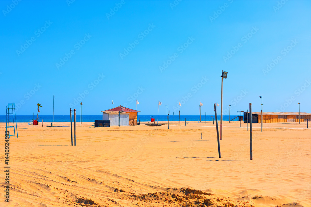 Wide sandy beach in Catania stretches under a clear sky, with calm sea in background and a lifeguard station near horizon. Volleyball nets, lifeguard tower, and scattered poles dot empty shoreline