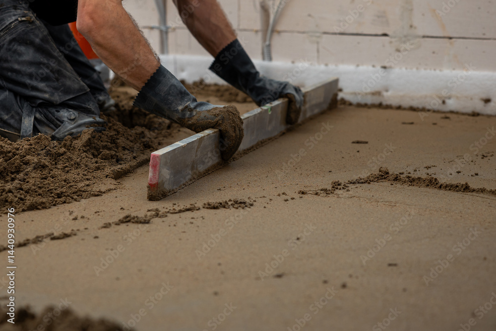 A worker kneels on wet concrete, leveling it with a straightedge tool. Underfloor heating pipes are visible, with unfinished walls and wiring around.
