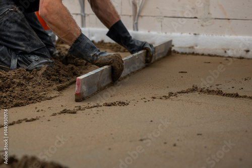 A worker kneels on wet concrete, leveling it with a straightedge tool. Underfloor heating pipes are visible, with unfinished walls and wiring around.