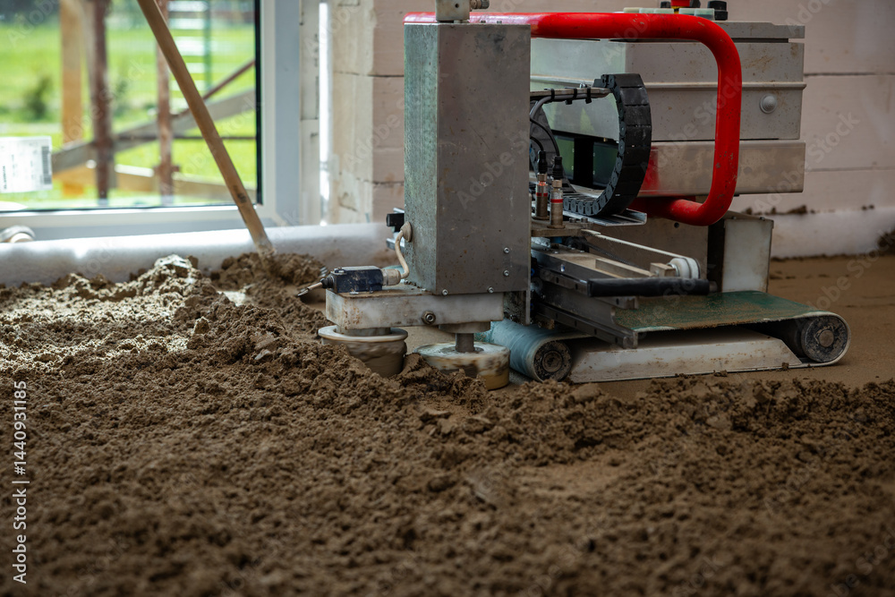 A concrete leveling machine with a red handle operates on wet material indoors. Natural light enters through a large window, revealing blurred green surroundings.