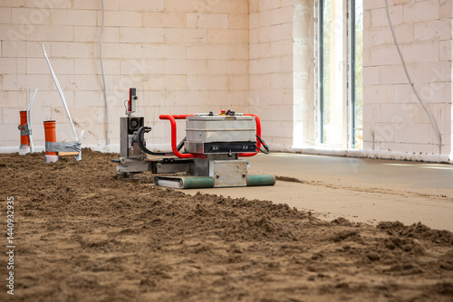 A construction site with a machine smoothing Estrich concrete over underfloor heating pipes. Unfinished white brick walls and large windows are visible.