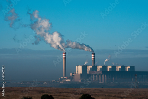The Kendal Power Station stands prominently in an open landscape, with tall chimneys emitting smoke under a clear blue sky; sharp focus highlights the industrial structure against the natural surround