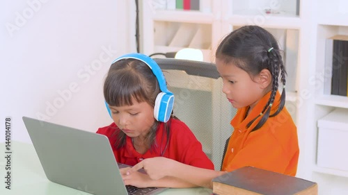 Two young asian girls collaborating on a laptop for online education, wearing headphones and glasses, with books stacked nearby, embodying remote learning and teamwork