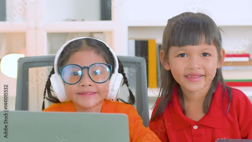 Two young asian girls collaborating on a laptop for online education, wearing headphones and glasses, with books stacked nearby, embodying remote learning and teamwork