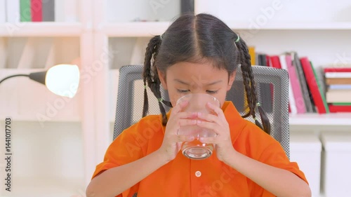 Healthy asian child enjoying a refreshing glass of pure water while sitting on a chair at home, emphasizing the importance of hydration and promoting a vibrant lifestyle