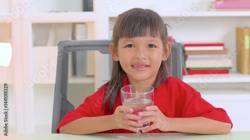 Healthy asian child enjoying a refreshing glass of pure water while sitting on a chair at home, emphasizing the importance of hydration and promoting a vibrant lifestyle