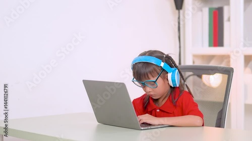 Asian girl studying online using laptop and wearing headphones sitting at desk in her room with bookshelf in background, homeschooling concept