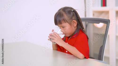 Healthy asian child enjoying a refreshing glass of pure water while sitting on a chair at home, emphasizing the importance of hydration and promoting a vibrant lifestyle