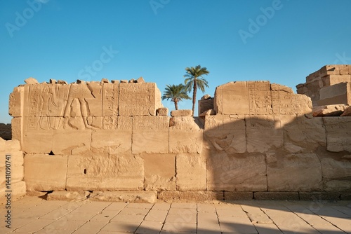 Luxor, Egypt - December 26 2023: Ruins of the Karnak Temple. Stone walls with carved drawings, hieroglyphs and palm trees with clear blue sky