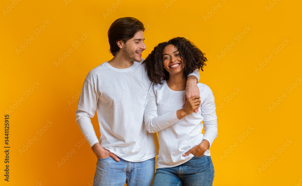 Portrait of happy multiracial couple hugging and posing together over yellow background in studio, romantic guy cuddling his afro girlfriend