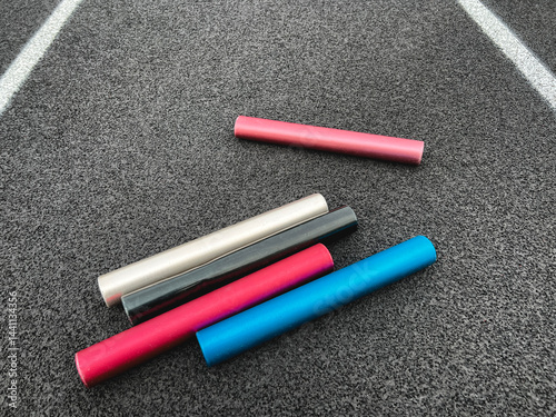 Relay Batons on a Running Track. Multi-colored aluminum relay batons rest on a black running track with white lane lines. Textured artificial rubber black surface.