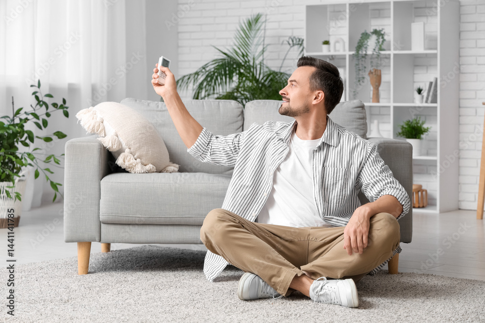 Handsome young man with air conditioner remote control sitting on floor in living room