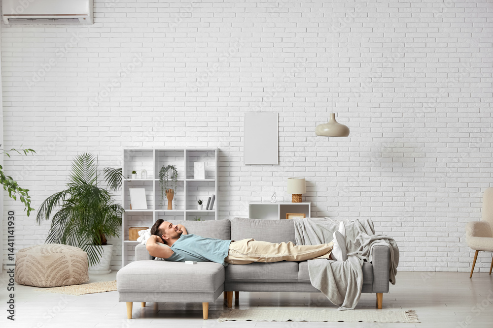 Handsome young man resting on sofa with operating air conditioner in living room