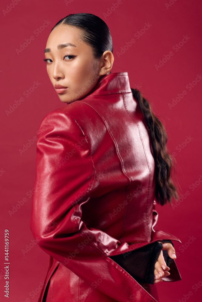 Fashionable asian woman in a red leather jacket posing with hands on hips against a red background, hair styled in a ponytail Fashionable asian woman in a red leather jacket posing with hands on hips against a red background, hair styled in a ponytail