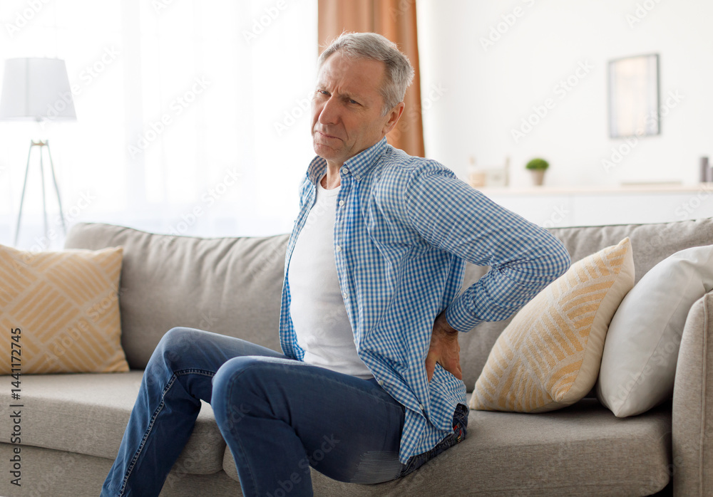 Backache And Health Problem Concept. Portrait of sick mature man feeling acute back pain, sitting on couch at home, person touching lower back aching area with hand, suffering from chronic illness Backache And Health Problem Concept. Portrait of sick mature man feeling acute back pain, sitting on couch at home, person touching lower back aching area with hand, suffering from chronic illness