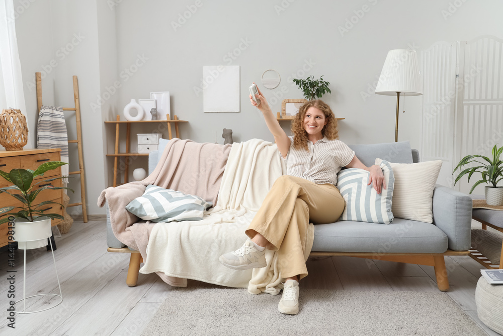 Beautiful smiling young woman with air conditioner remote control sitting on sofa in living room
