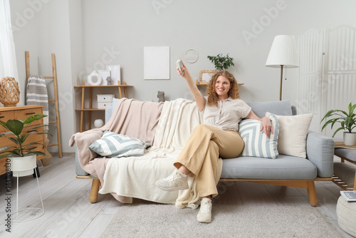 Obraz na płótnie Beautiful smiling young woman with air conditioner remote control sitting on sofa in living room w salonie