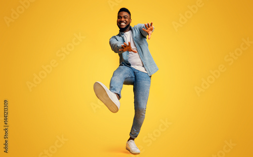 Funny african american guy dancing and extending his hands to camera over yellow studio background, low-angle view