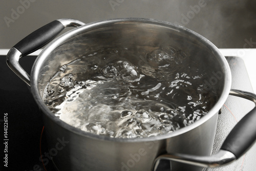 Cooking pot with boiling water and stove on white table against grey background, closeup