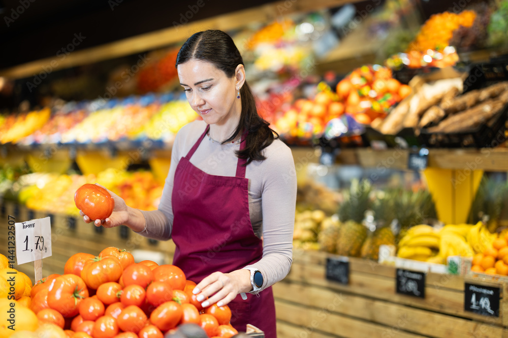 Obraz premium Glad middle-aged female marketer setting tomatoes in market basket in big greengrocery