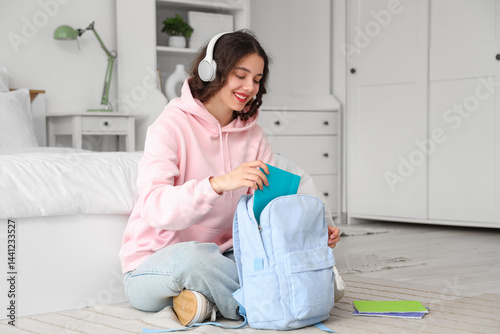 Female Indian student with headphones taking copybook from backpack in bedroom
