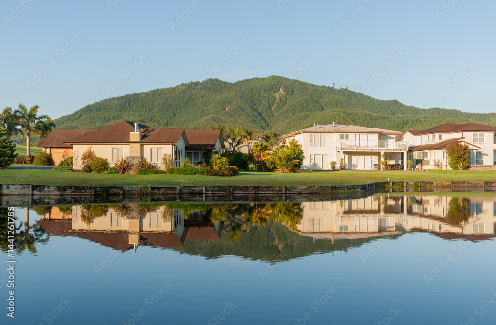 Idyllic residential home reflections in calm waterway foreground and green hill backdrop under blue sky.
