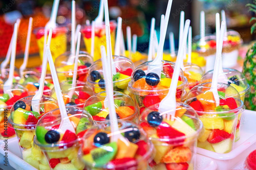 Colorful cups of fresh mixed fruit, including strawberries, kiwi, melon, and blueberries, neatly arranged with plastic forks. A vibrant, healthy snack option at a market or food stand